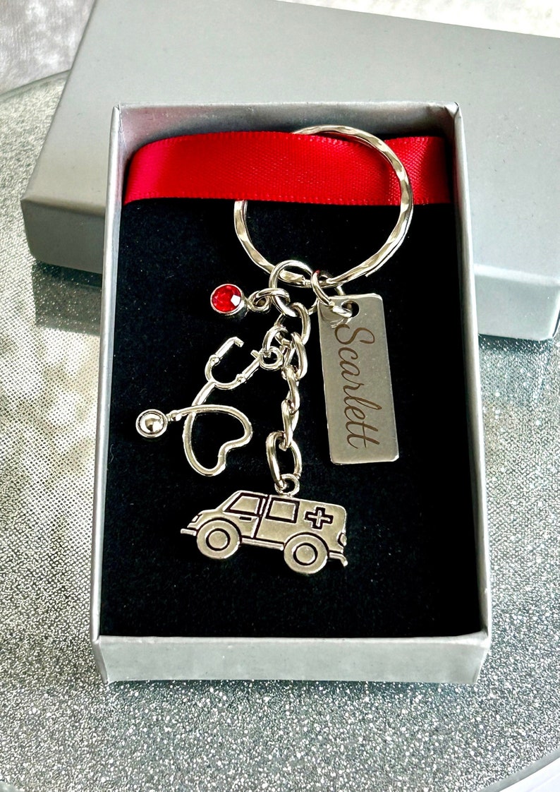May include: A silver keychain with a red rhinestone, a stethoscope, a heart, and a silver ambulance charm. The keychain also has a silver tag with the name "Scarlett" engraved on it.