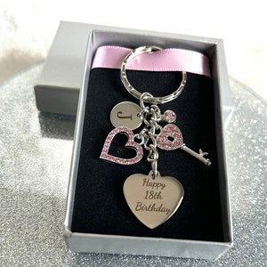 May include: A silver keyring with pink rhinestones in the shape of hearts and a key. The keyring also has a round charm with the letter J engraved on it. The keyring is in a grey box with a pink ribbon. The heart charm has the text "Happy 18th Birthday" engraved on it.