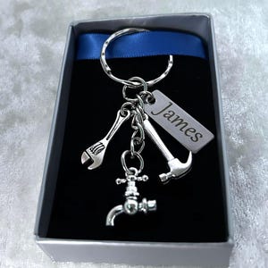 May include: A silver keyring with charms shaped like a wrench, hammer, and tap. The keyring includes a tag engraved with the name "James". Presented in a gift box with a blue ribbon.