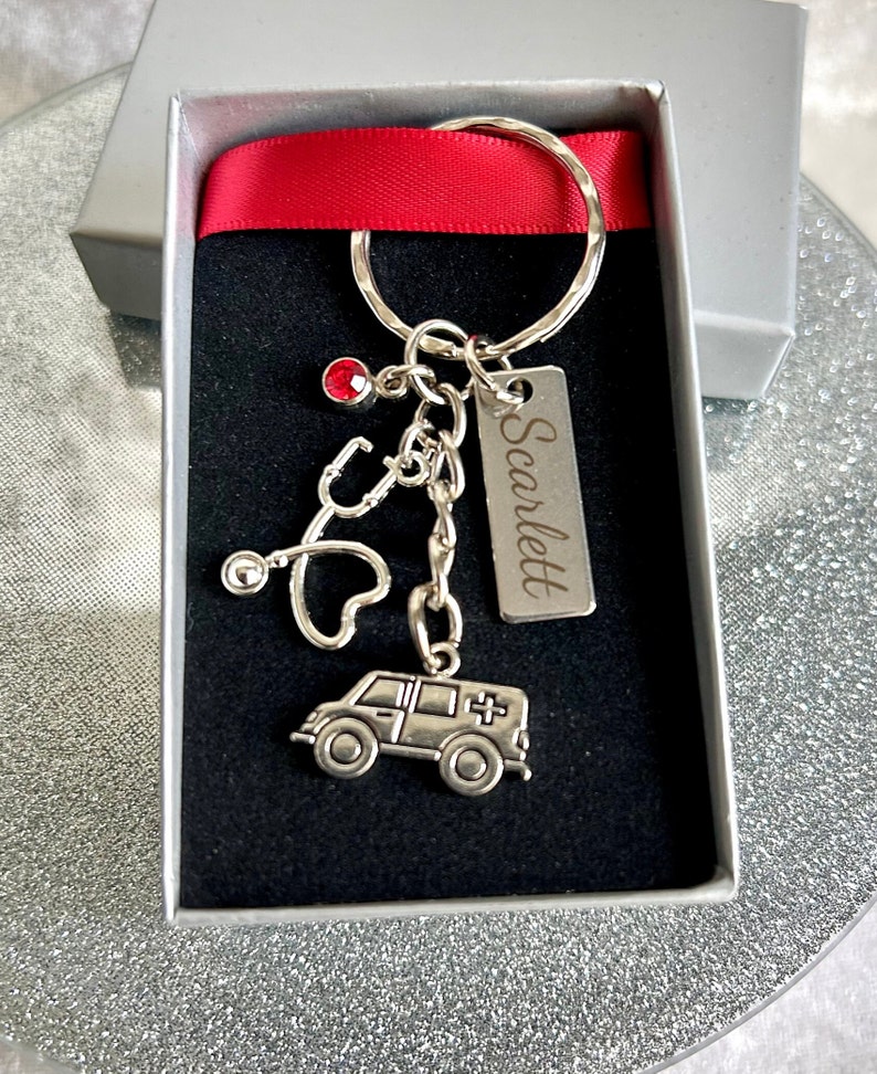 May include: A silver keychain with a red rhinestone, a stethoscope, a heart, and a small ambulance charm. The keychain also has a silver tag with the name "Scarlett" engraved on it.
