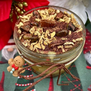 May include: A clear glass jar candle with a brown lid and a gingerbread man charm. The candle is filled with a brown wax and topped with brown glitter and small brown stones.