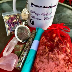 May include: A white mug with the text "Dreamer Apothecary Stay Wild Moon Child" on it. The mug is filled with tea and has a tea infuser with a heart charm hanging from it. There is a small bag of loose leaf tea, a glass vial of herbs, a heart-shaped pink crystal, a green and red crystal, a clear crystal, a red pouch with a white and pink mixture inside, and a blue and turquoise fabric wrapped around the vial of herbs.
