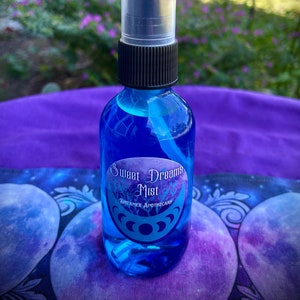 May include: A blue glass bottle with a black spray top and a white label that reads "Sweet Dreams Mist" with a moon and stars design. The bottle is sitting on a purple and blue fabric with a moon and stars design.