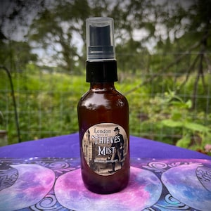 May include: A brown glass bottle with a black spray top and a white label that reads "London Thieves Oil Thieves Mist Dreamer Apothecary". The bottle is sitting on a purple and blue fabric with a moon pattern.