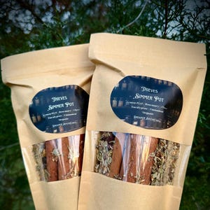 May include: Two brown paper bags with clear plastic windows containing Thieves Simmer Pot blends. The bags have labels with the text "Thieves Simmer Pot" and a list of ingredients: Lemon Peel, Rosemary, Clove, Eucalyptus, Cinnamon. The text "Organic" is also on the label. The bags are labeled "Dreamer Apothecary."