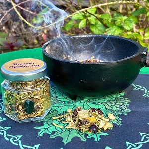 May include: A glass jar labeled "Dreamer Apothecary" filled with dried herbs and flowers. The jar is sitting on a green and black patterned cloth. A black cauldron with smoke rising from it is in the background.