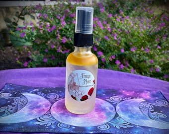 May include: A clear glass bottle with a gold liquid inside. The bottle has a black spray top and a white label that says "Freyja Mist" and "Dreamer Apothecary". The bottle is sitting on a purple fabric with a design of three moons.