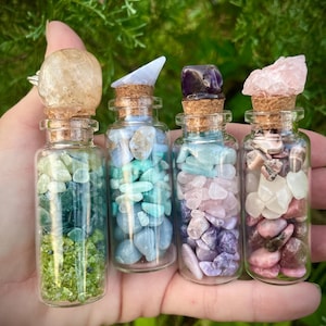 May include: Four small glass bottles filled with various colorful crystals and topped with cork stoppers. The bottles are held in a hand against a green leafy background.