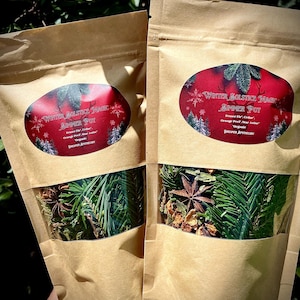 May include: Two brown paper bags with clear windows showing dried herbs and spices inside. The bags are labeled "Winter Solstice Magic Simmer Pot" with the ingredients listed as "Fraser Fir, Cedar, Orange Peel, Star Anise" and "Organic" with the Dreamer Apothecary logo.