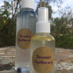 May include: Two clear glass spray bottles with white tops and gold labels that say "Dreamer Apothecary". The bottles are sitting on a wooden surface.
