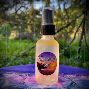 May include: A clear glass bottle with a black spray top and a round label that says "Florida Sunset Mist" with a sunset graphic. The label also says "Dreamer Apothecary".