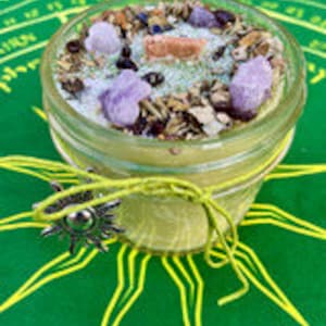 May include: A yellow candle in a glass jar with a green, brown, and white herbal blend on top. The jar is tied with a yellow ribbon and a silver charm.