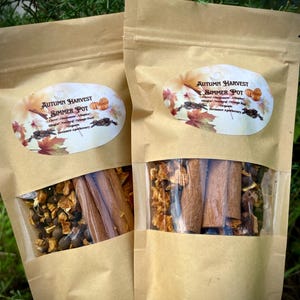 Może przedstawiać: Dwie brązowe papierowe torby z przezroczystymi okienkami z plastiku, każda zawierająca mieszankę suszonych przypraw i skórki pomarańczowej. Torby są oznaczone "Autumn Harvest Simmer Pot" z listą składników i "Dreamer Apothecary".