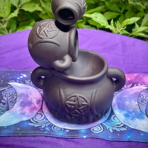 Op de afbeelding: Een bruine keramische wierookbrander met een pentagram-ontwerp. De brander is gevormd als een ketel en heeft een kleinere, ronde bovenkant. De brander staat op een paarse stof met een maan- en sterrendesign.