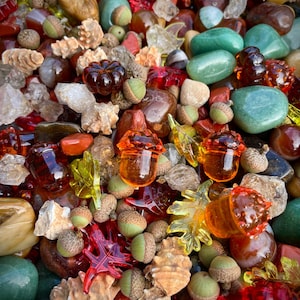 May include: A collection of decorative glass acorns, leaves, and pumpkins in shades of amber, red, and green. The assortment includes natural stones and seashells, creating a fall-themed display.
