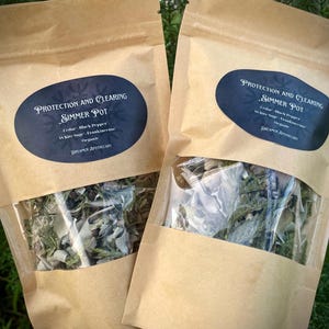 Peut inclure: Deux sacs en papier kraft avec des fenêtres transparentes montrant des herbes séchées à l'intérieur. Les sacs sont étiquetés "Protection and Clearing Simmer Pot" avec une liste d'ingrédients : Cèdre, Poivre noir, Sauge blanche, Encens, Bio. Les sacs sont de Dreamer Apothecary.