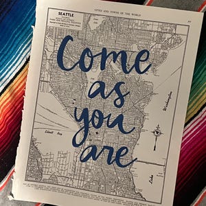 May include: A vintage map of Seattle with the words "Come as you are" in blue script. The map is detailed with city streets and water features, and is set against a colorful striped background.
