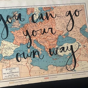 May include: Vintage map of Europe and the Mediterranean with the words "you can go your own way" in elegant black script. The map features a color palette of muted blues, oranges, and tans, with country borders clearly defined.