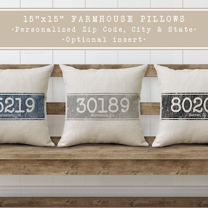 Op de afbeelding: Drie vierkante farmhouse-stijl kussens van 38 cm met gepersonaliseerde postcode, stad en staat. De kussens zijn gemaakt van natuurlijke linnen stof met een verweerde look. De postcodes zijn 45219, 30189 en 80202.