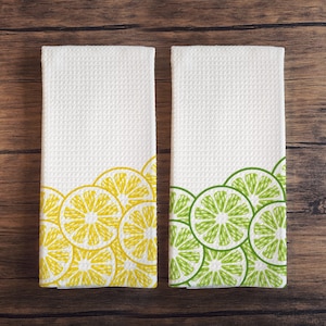 May include: Two white kitchen towels with a border of yellow lemon slices on one towel and green lime slices on the other towel.