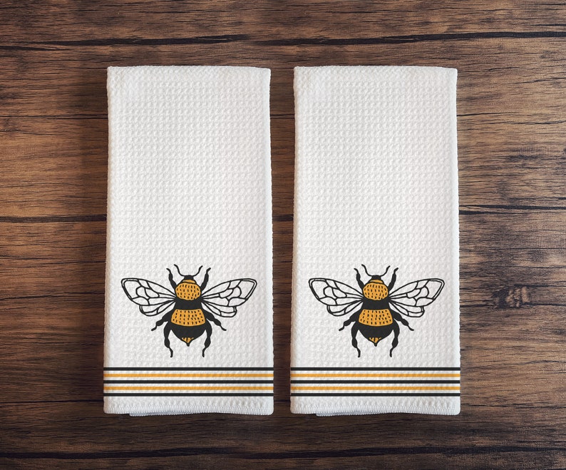 May include: Two white kitchen towels with black and yellow bumble bee designs. The towels have black and yellow stripes at the bottom.