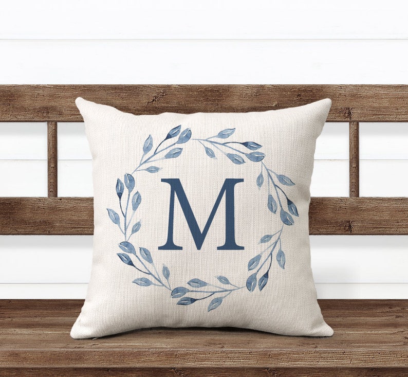 May include: A white throw pillow with a blue watercolor wreath and the letter "M" in the center. The pillow is sitting on a wooden bench.