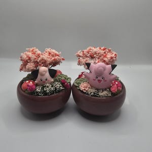 May include: Two matching decorative planters, each featuring a cartoon character with pink skin, black ears, and a smiling face. The planters are filled with greenery, pink flowers, and a small tree with pink and red blossoms.