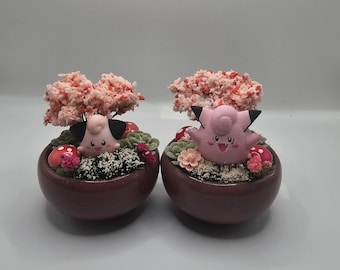 Könnte beinhalten: Zwei passende dekorative Pflanzgefäße, jeweils mit einer Cartoonfigur mit rosa Haut, schwarzen Ohren und einem lächelnden Gesicht. Die Pflanzgefäße sind mit Grün, rosa Blumen und einem kleinen Baum mit rosa und roten Blüten gefüllt.