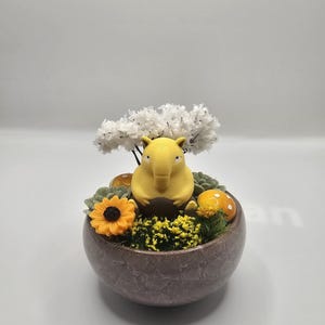 Puede incluir: Un jardín en miniatura decorativo en una maceta de cerámica marrón. Presenta una figura de elefante amarillo, un árbol blanco, un girasol naranja y setas amarillas. El jardín está lleno de follaje verde y amarillo.
