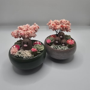 May include: Two decorative ceramic bowls, one dark green and one gray, each containing miniature bonsai-style arrangements. The arrangements feature small brown figures, pink and red mushroom accents, and miniature pink flowering trees.