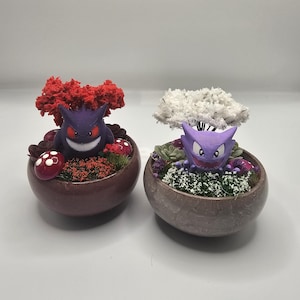 Puede incluir: Dos jardineras decorativas con personajes de dibujos animados morados con ojos rojos y caras sonrientes. Una jardinera tiene un árbol rojo, la otra un árbol blanco. Ambas jardineras tienen follaje verde y rojo y están en macetas ovaladas marrones.
