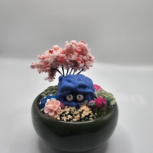 Könnte beinhalten: Ein skurriles Miniatur-Bonsai-Arrangement in einem dunkelgrünen Keramiktopf. Es zeigt eine blaue Kreatur mit großen Augen, einen rosa blühenden Baum, bunte Dekorationselemente und künstliches Grün.