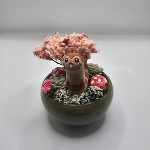 Könnte beinhalten: Ein dekorativer Keramiktopf mit einem skurrilen Design. Er zeigt eine cartoonartige Kreatur, einen Miniatur-Blütenbaum in Rosa, Sukkulenten und rote und weiße Pilzakzente. Der Topf hat eine dunkelgrüne Farbe.