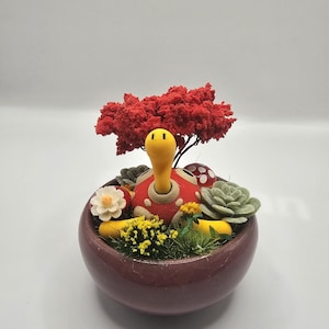 Könnte beinhalten: Ein handgefertigter Keramik-Blumentopf mit einer schildkrötenartigen Figur. Der Topf ist mit Miniaturpflanzen, Blumen und einem kleinen roten Baum gefüllt. Der Topf hat eine tief burgunderrote Farbe. Die Schildkröte hat einen gelben Kopf und einen roten Panzer.