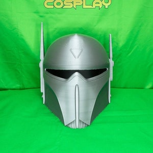May include: A 3D printed silver helmet with a visor and two antennas. The helmet is on a green background. The text "PRINTING COSPLAY" is visible at the top of the image.