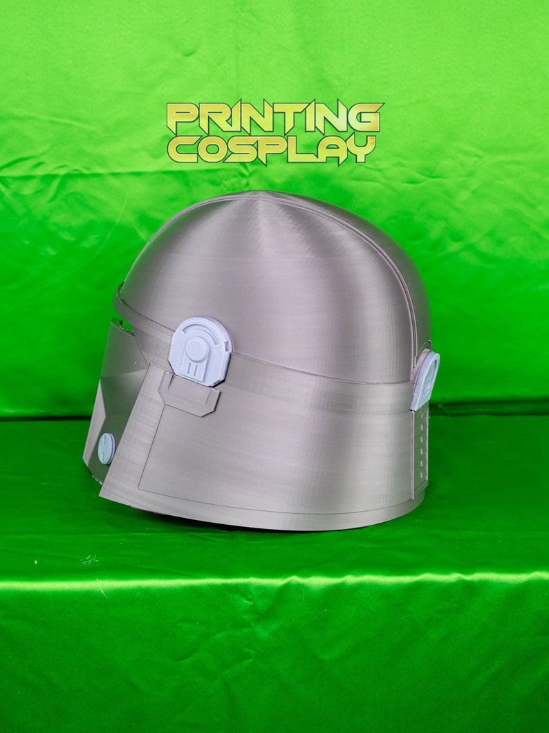 May include: A silver-colored helmet with a visor and ear pieces, likely for cosplay or costume use. The helmet has a smooth, metallic finish and is set against a bright green background. The text "PRINTING COSPLAY" is visible at the top.