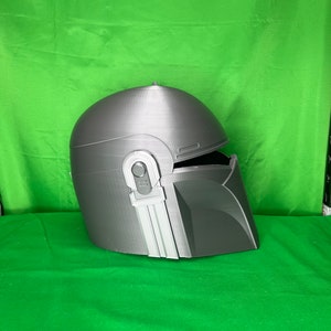 May include: A gray 3D printed helmet with a visor and a textured surface. The helmet has a design that resembles a Mandalorian helmet.