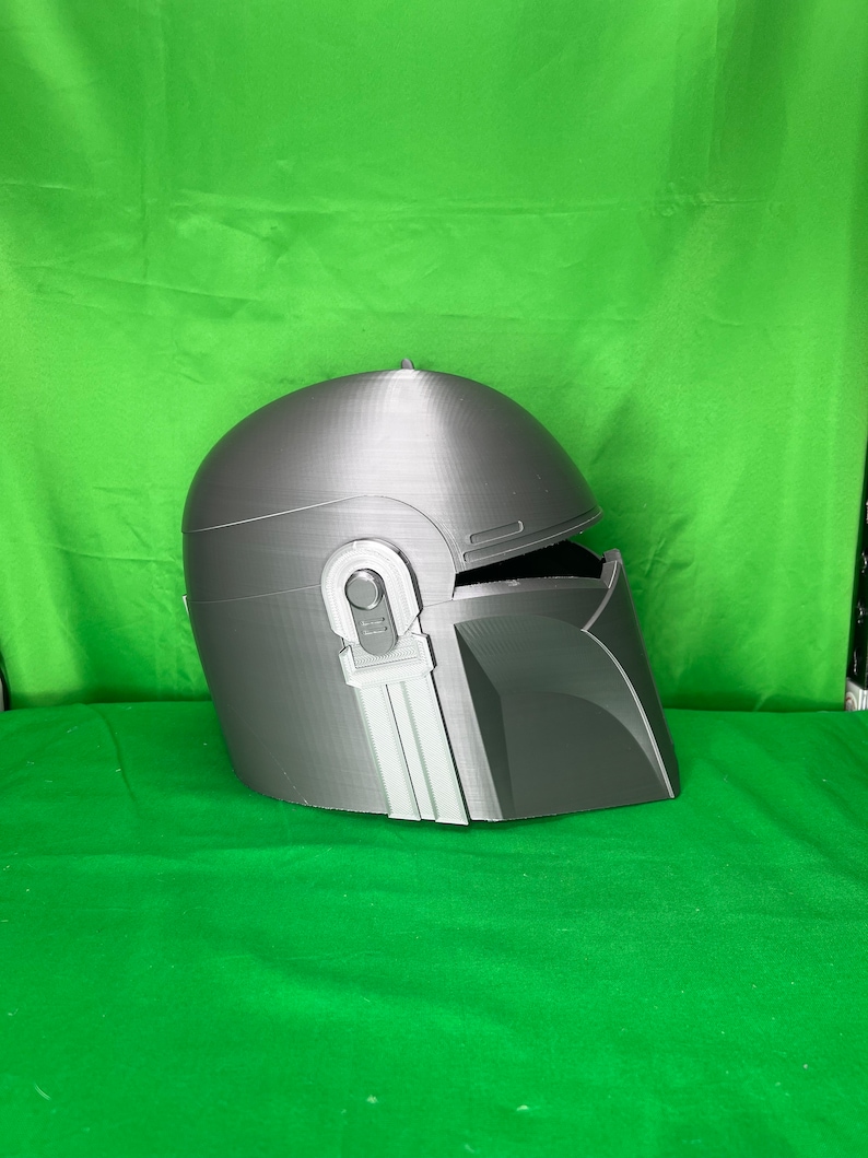 May include: A gray 3D printed helmet with a visor and a textured surface. The helmet has a design that resembles a Mandalorian helmet.