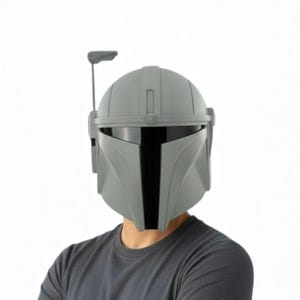 May include: A grey Mandalorian helmet with a black visor and a vertical T-shaped opening. The helmet has a rounded top and a small antenna on the left side. The person is wearing a grey t-shirt.