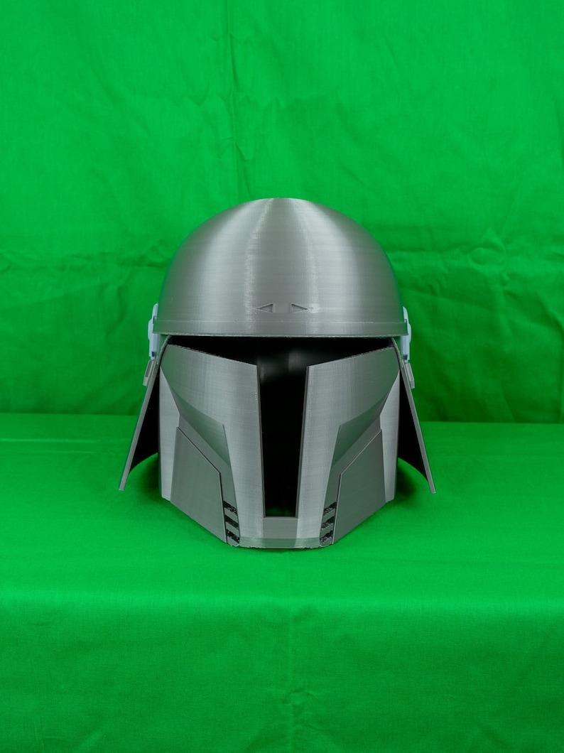 May include: A silver-coloured helmet with a textured surface. The helmet has a visor and a raised design on the top.
