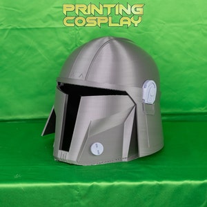 May include: A silver Mandalorian helmet with a detailed design, featuring a visor and various angular panels. The helmet is displayed on a green surface, with the text "PRINTING COSPLAY" in gold lettering.