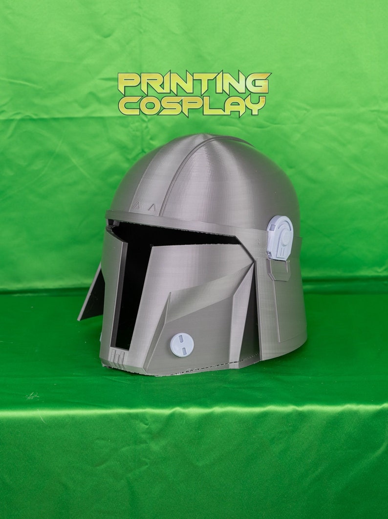 May include: A silver Mandalorian helmet with a detailed design, featuring a visor and various angular panels. The helmet is displayed on a green surface, with the text "PRINTING COSPLAY" in gold lettering.