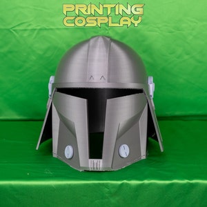 May include: A gray Mandalorian helmet with a rounded top and angular features. The helmet has a visor opening and two circular details on the lower front. The text "PRINTING COSPLAY" is displayed in gold letters above the helmet.