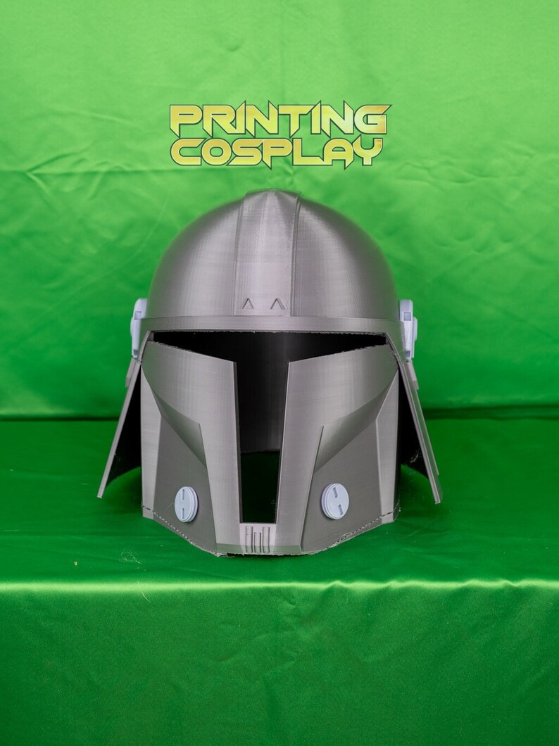 May include: A gray Mandalorian helmet with a rounded top and angular features. The helmet has a visor opening and two circular details on the lower front. The text "PRINTING COSPLAY" is displayed in gold letters above the helmet.