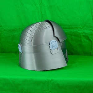 May include: A grey 3D printed helmet with a textured surface and a raised design. The helmet has a hinged section at the back and two small, light grey details on the side.