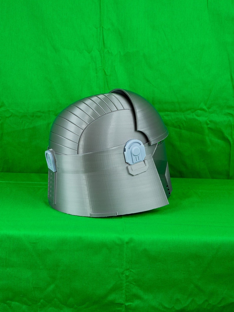 May include: A grey 3D printed helmet with a textured surface and a raised design. The helmet has a hinged section at the back and two small, light grey details on the side.