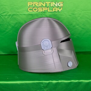 May include: A silver-colored Mandalorian helmet with a detailed design, featuring a visor and various attachments. The helmet is displayed against a green backdrop, with the text "PRINTING COSPLAY" in gold letters.