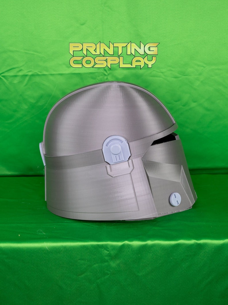 May include: A silver-colored Mandalorian helmet with a detailed design, featuring a visor and various attachments. The helmet is displayed against a green backdrop, with the text "PRINTING COSPLAY" in gold letters.