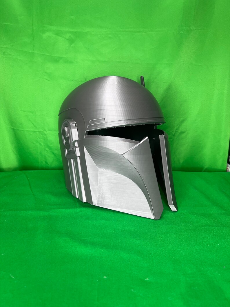 May include: A gray 3D printed helmet with a textured surface and a raised design on the front. The helmet has a visor and a small antenna on top.