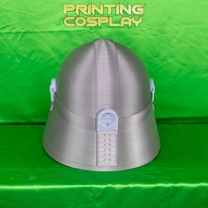 May include: A silver-colored cosplay helmet with a rounded top and a flared base. The helmet has white accents and a rectangular design on the front. The text "PRINTING COSPLAY" is displayed in a yellow-green font.